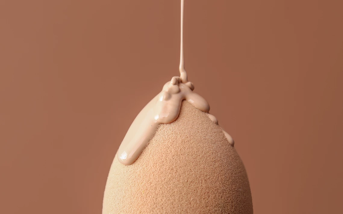 Ein Make-up-Schwamm in warmem Beige, auf den flüssige Foundation tropft, vor einem braunen Hintergrund.